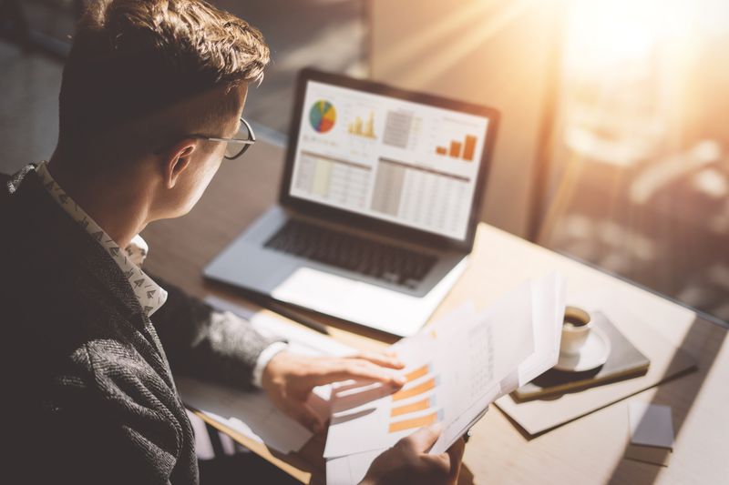 Young market analyst looking at laptop and papers with financial information and an espresso on his desk