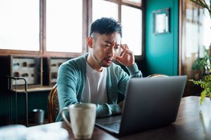 Worried young Asian man with his hand on head, using laptop computer at home, looking concerned and stressed out
