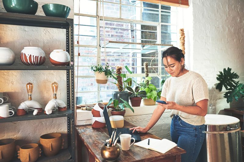 A person using a phone while standing by a desk in a room with pottery items and potted plants