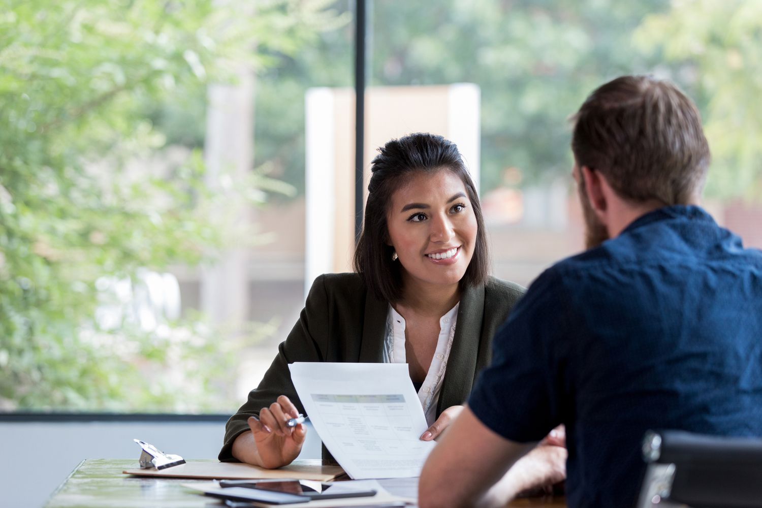 The image shows a woman sitting at a table, holding up a document and smiling while talking to a man seated across from her. The setting appears to be an office or meeting space, with large windows in the background offering a view of greenery outside. The woman seems to be explaining or discussing something from the document with the man, creating a professional and friendly interaction.
