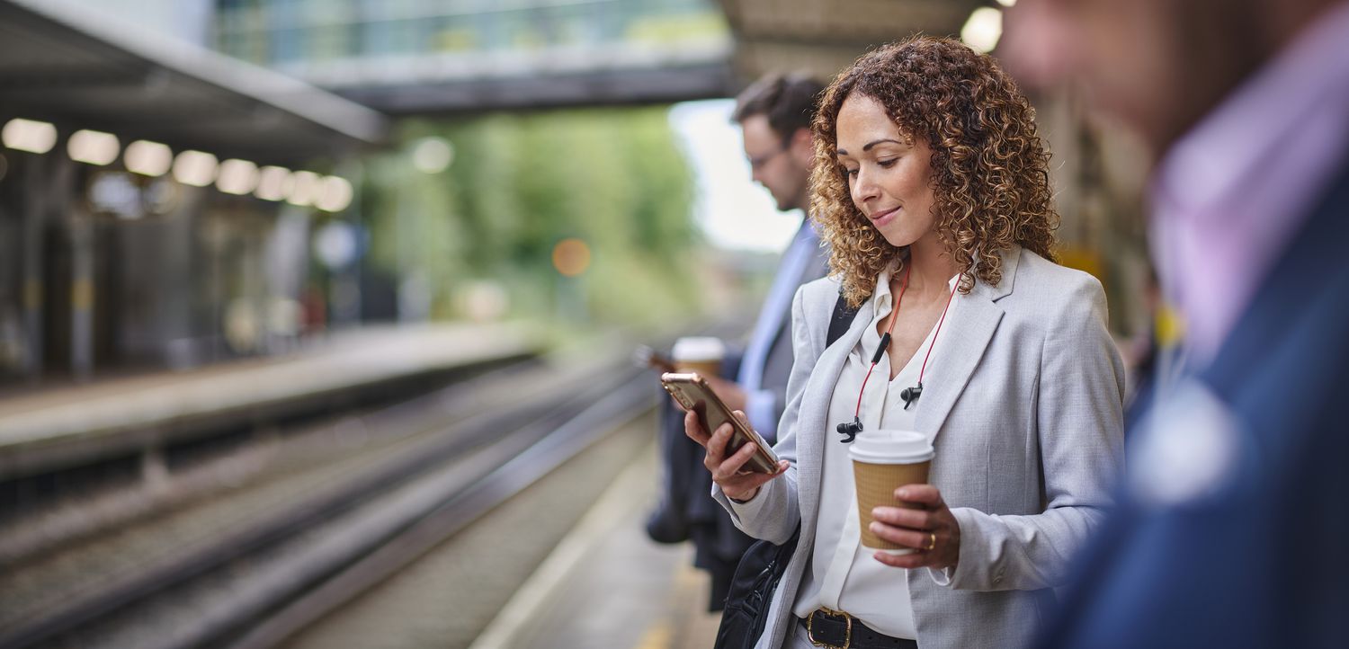 Woman on a train platfor reading her phone while holding a cup of coffee. 