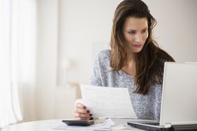 Woman looking at a laptop and holding a piece of paper as a calculator sits on her table.