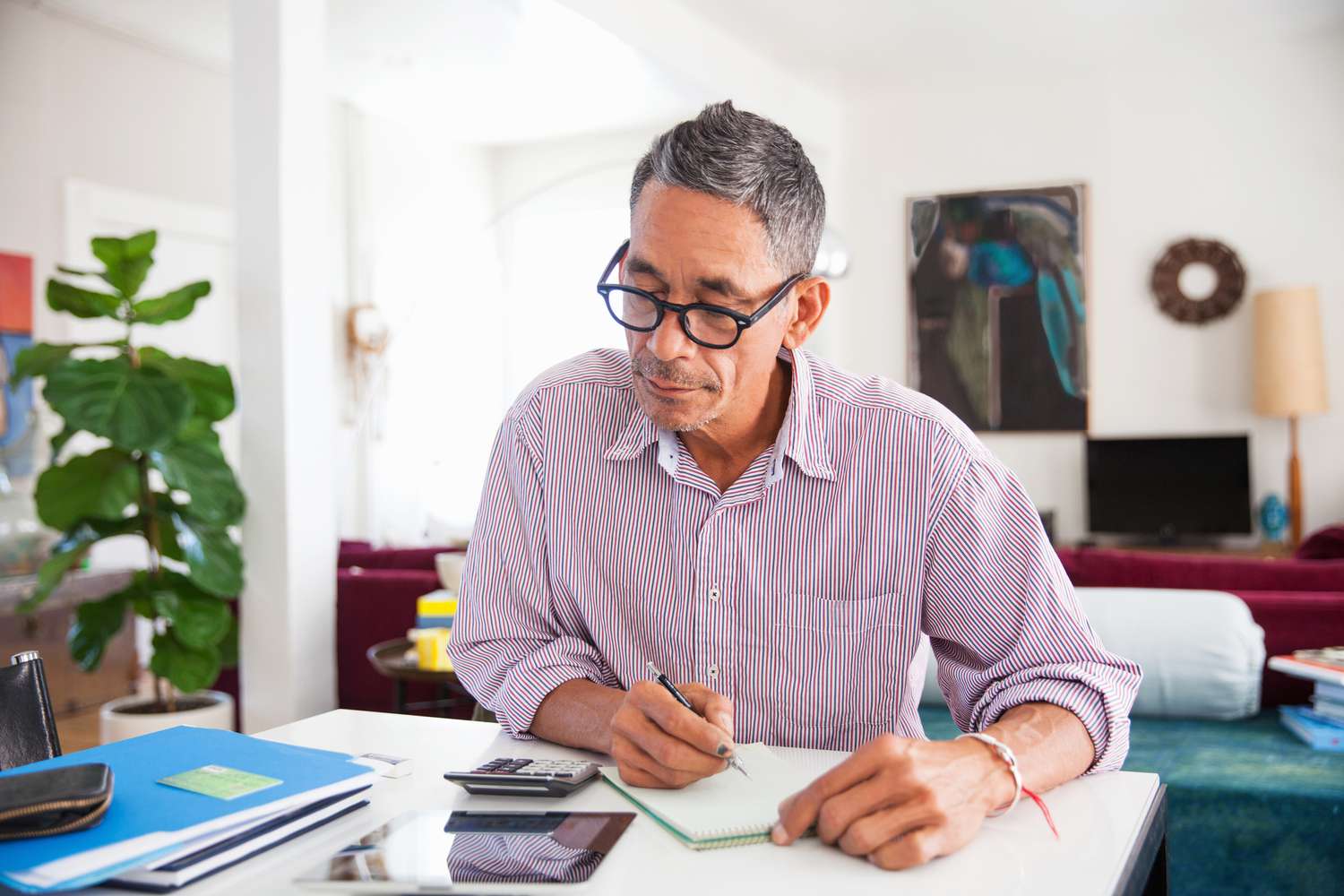 A man with short gray hair and glasses sits at a desk in a bright, cozy room. He is focused on writing in a small notepad, with a calculator, tablet, and papers spread out in front of him. The background features a potted plant, artwork, and comfortable furnishings.