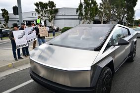 Protestors stand between a Tesla dealership and a Tesla Cybertruck driving on the street.