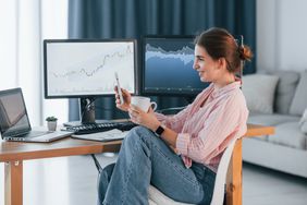 Crypto trader in casual clothes works at home office with a cup of drink and smartphone in hands.