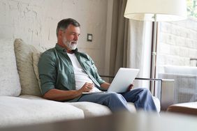 Man in his 50s or 60s sitting on his living room couch and studying something on his laptop