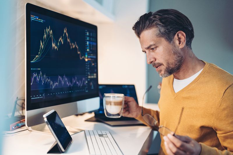 A retail day trader holds a cup of coffee as they work at home reading data from multiple screens showing stock charts.