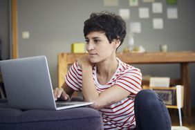 A woman in a red and white striped t-shirt sits on the floor against a couch with a laptop