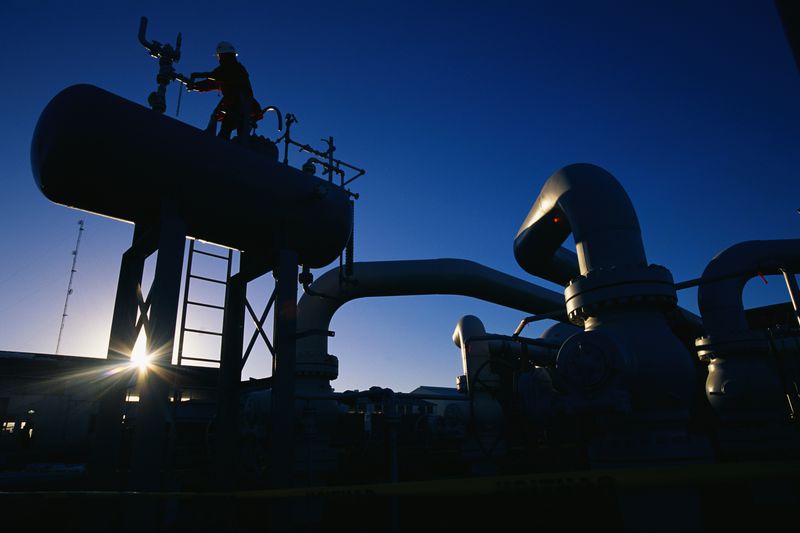 A natural gas production facility is silhouetted against the rising sun as a worker turns a valve on top of a large tank.