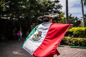 A person wearing a sombrero holds the Mexican flag in a park setting