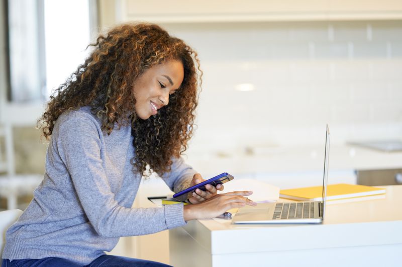 Woman sits at kitchen counter looking at laptop and cell phone