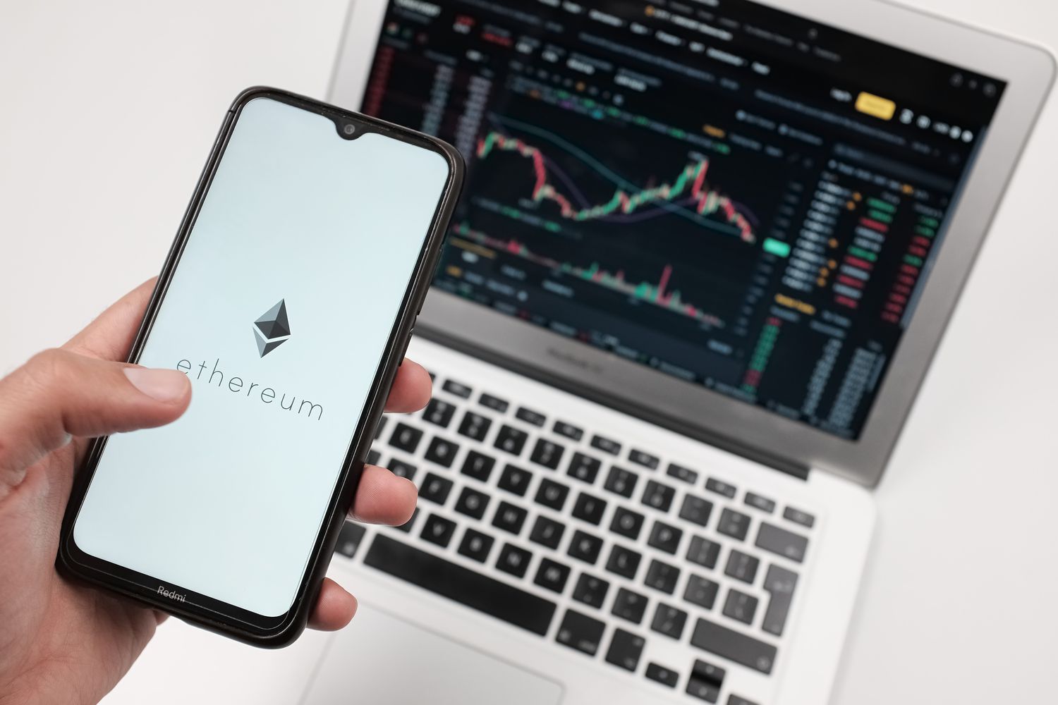 A person holds a cell phone with an Ethereum logo, while a laptop displaying stock charts appears in the background.