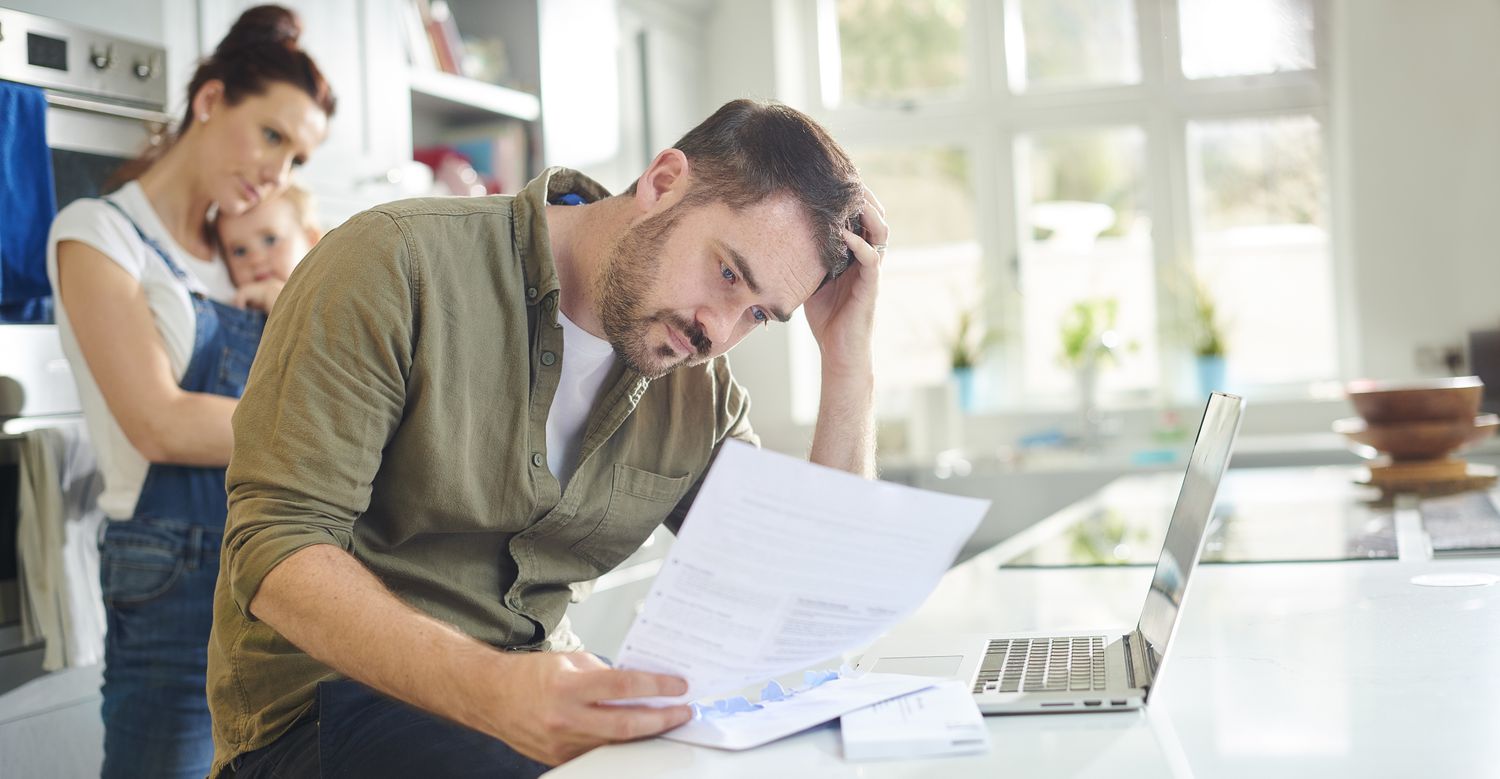 A person looks at bills with a mother and child in the background. All are in a kitchen.