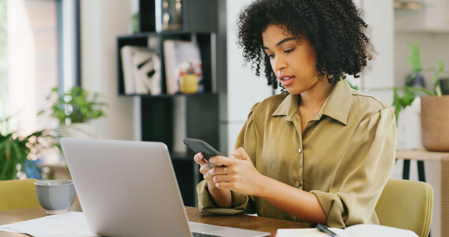 Someone using a smartphone and a laptop at a kitchen table.
