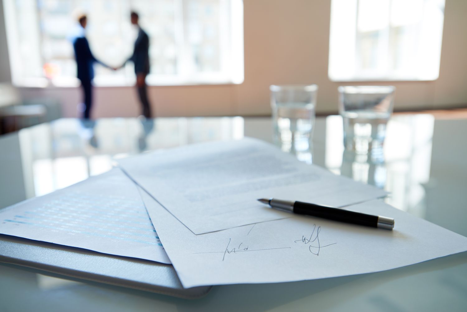Two people standing and shaking hands with a signed contract in the foreground