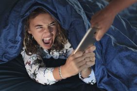 A hand reaches into the frame to take an iPhone away from an upset teenager under their covers in bed.