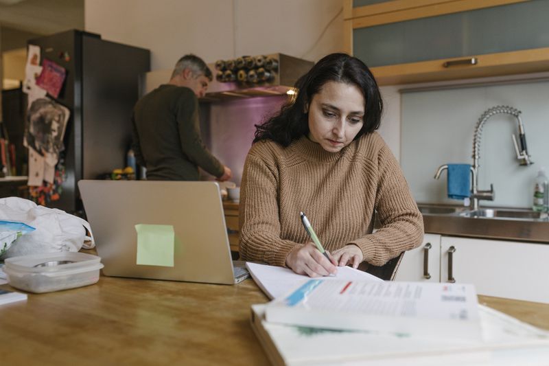 Mature woman working on the paper for her loan in the kitchen, husband cooking behind