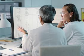 Two women wearing lab coats look at data on a computer screen in a hospital setting