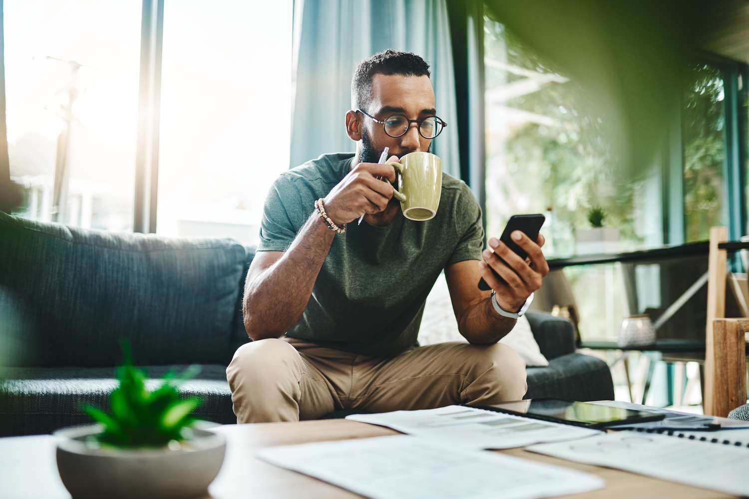 A man sits on his couch looking at his phone while drinking from a mug