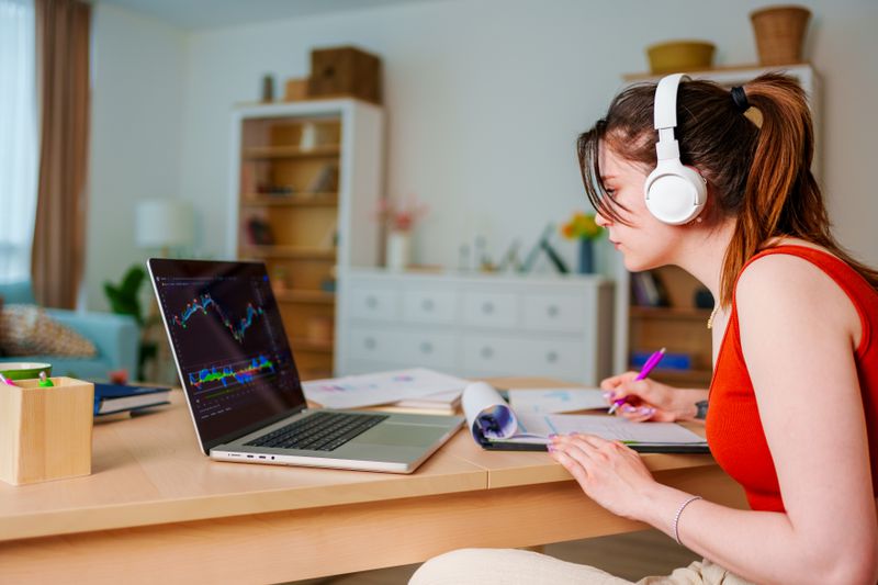 Young forex trader listening to the news while trading on laptop and also taking notes into trading journal. 