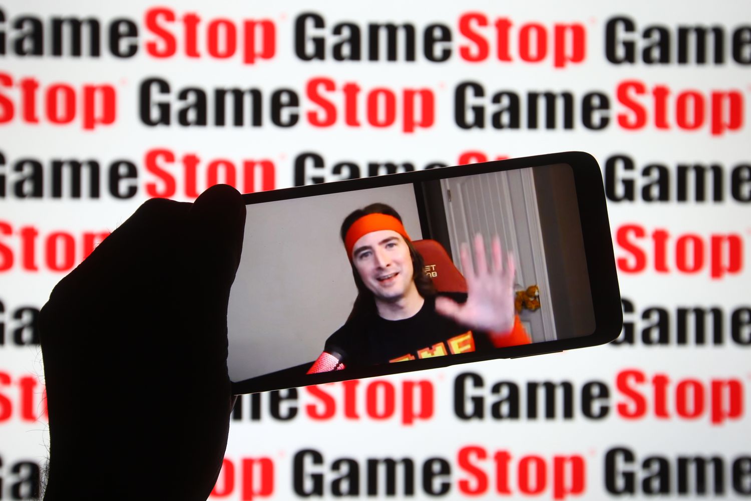 Keith Gill, known on Reddit as the pseudonym "Roaring Kitty," seen on a fragment of a YouTube video displayed on a smartphone screen in front of the GameStop logo