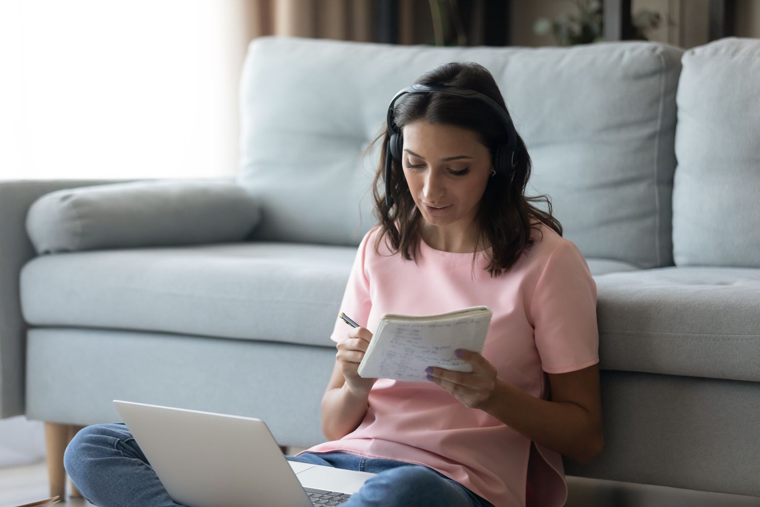 Woman sitting on the floor while making notes while looking at documents on laptop.