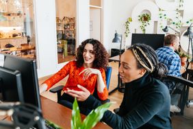 A transgender woman smiling while working at a computer desk with one of her colleagues