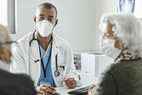 Doctor discussing with elderly couple during COVID-19 outbreak. Male healthcare worker is explaining senior man and woman while sitting at desk in clinic. They are in protective face masks.