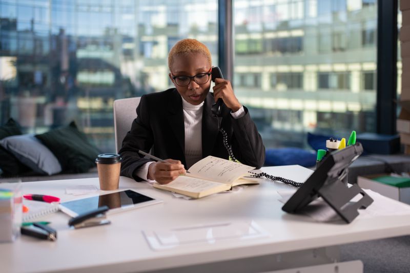 Woman sitting at a desk in a office on a phone call while taking notes.