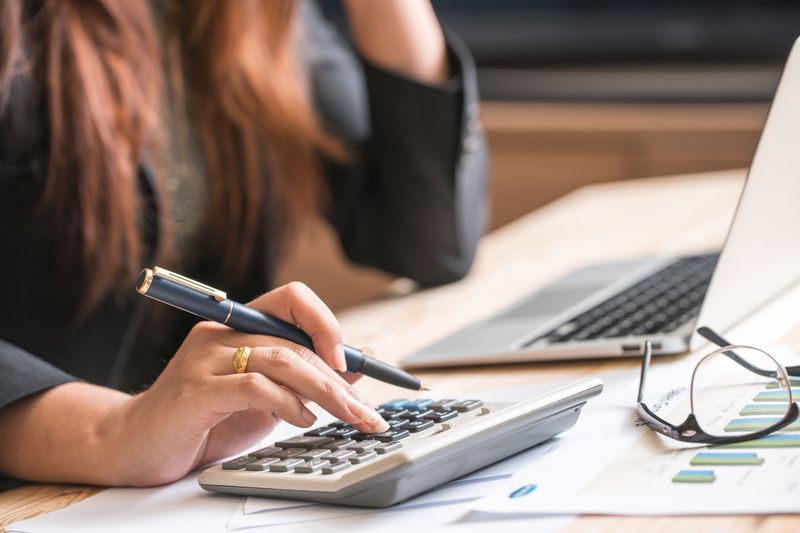Woman's hand on a calculator with a pen in front of a laptop and a pair of glasses