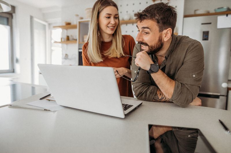 A happy couple stand at their kitchen table, looking at a laptop.