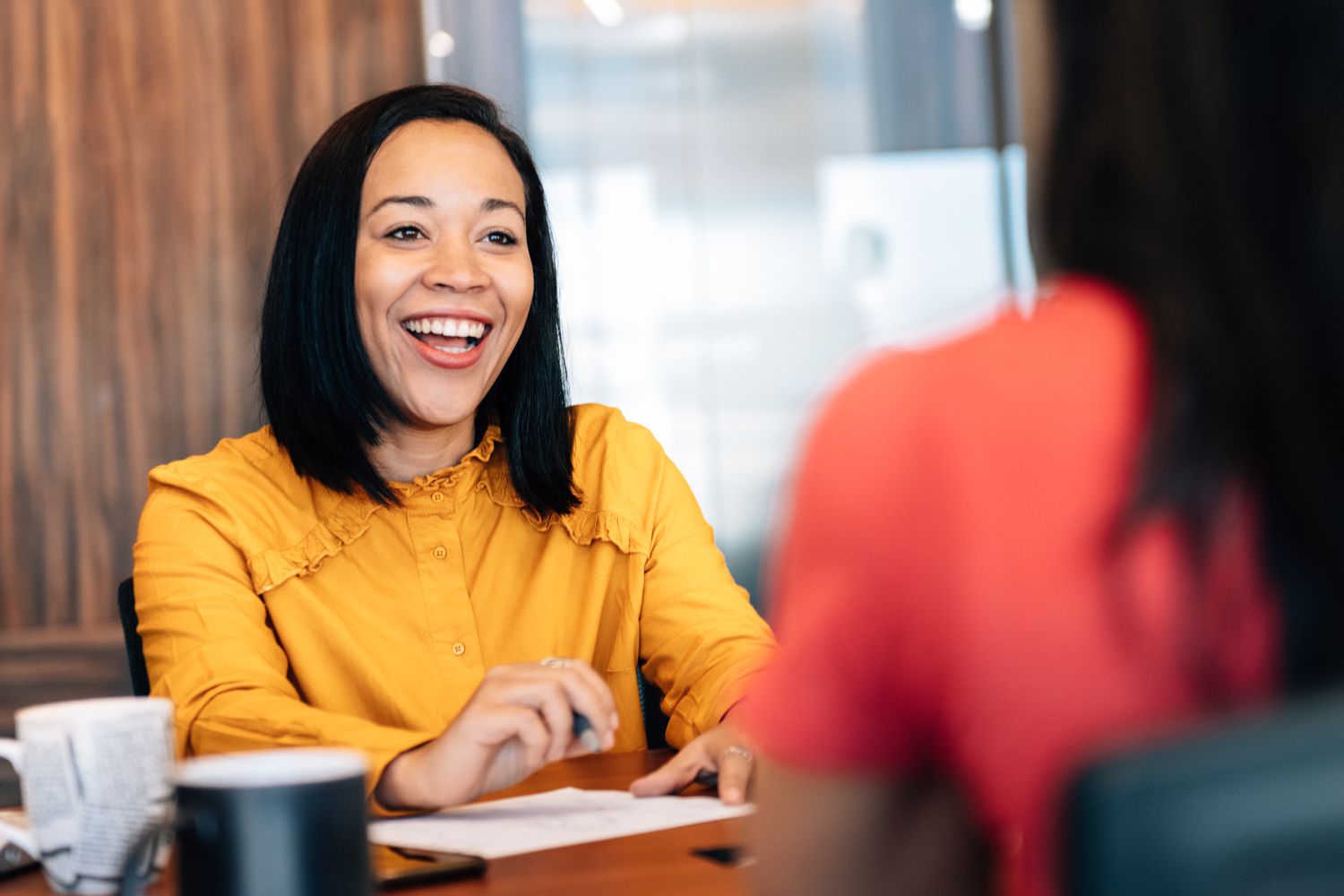woman smiling as she sits across a desk from someone else, holding paper.