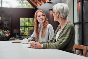A younger adult woman talks with an older adult woman at a table with coffee.