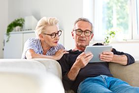 Middle-aged man reclining on a white couch holding a tablet. His wife squatting behind the couch is looking over his shoulder at the tablet. 