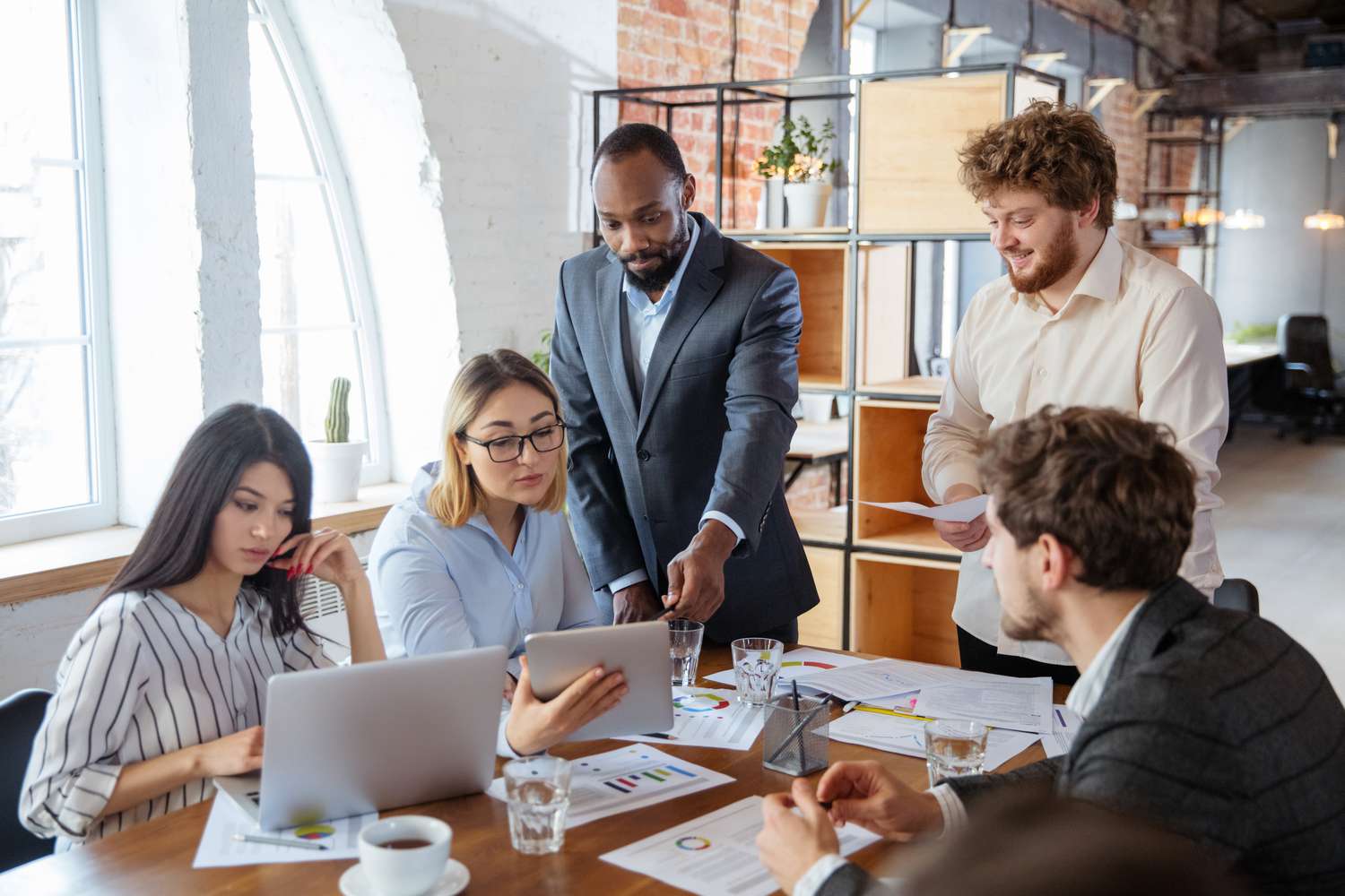 A meeting of co-workers use financial data and laptops to find places to offset the cost of a new office equipment upgrade using a zero-cost strategy.