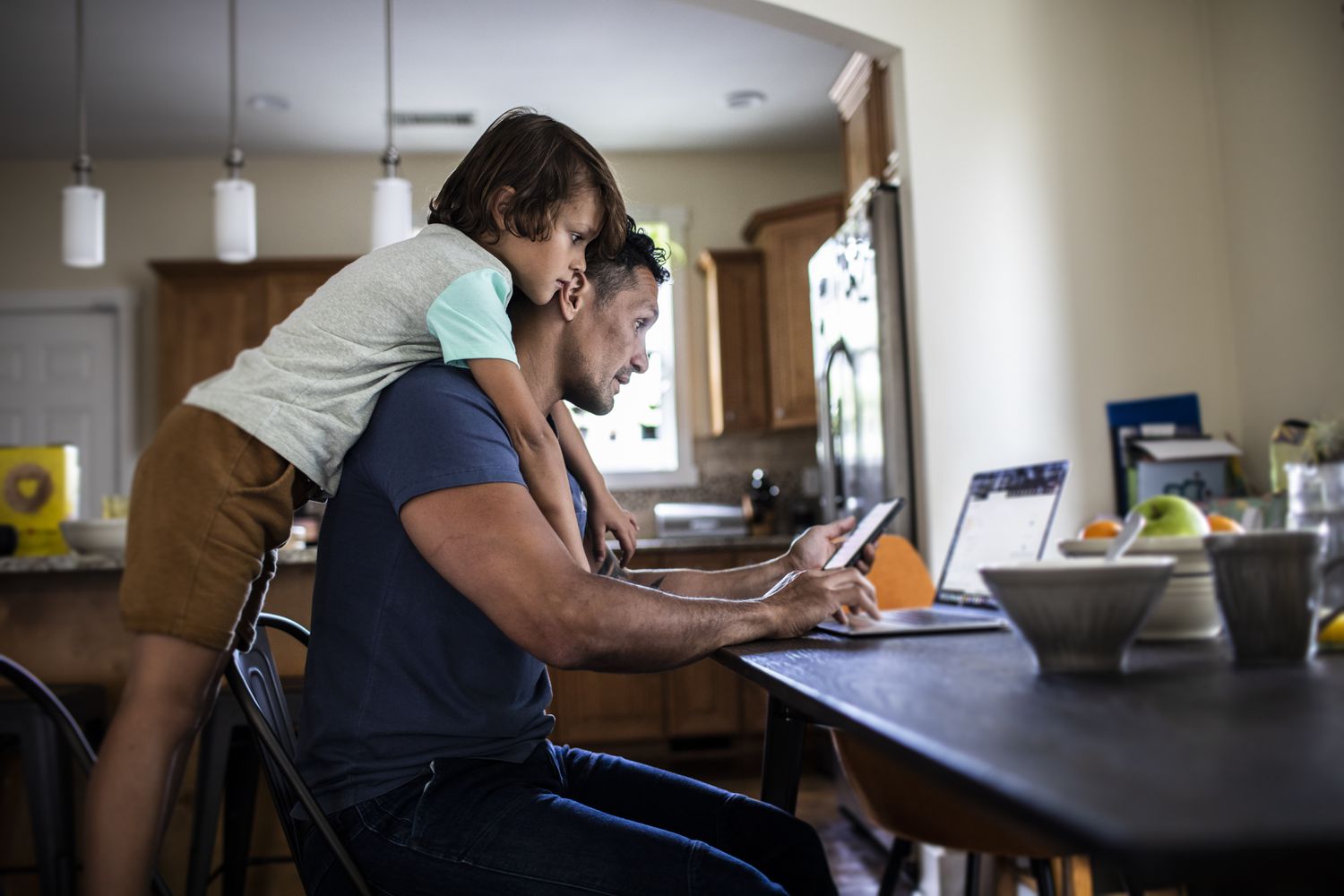 man looking at phone and laptop while child looks over his shoulder