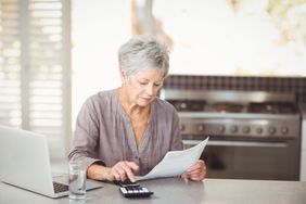 Older woman sitting in her kitchen and looking at a mortgage document, a calculator and a laptop