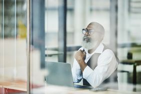 Medium shot view through window of senior businessman looking out window while working on laptop in coworking office space