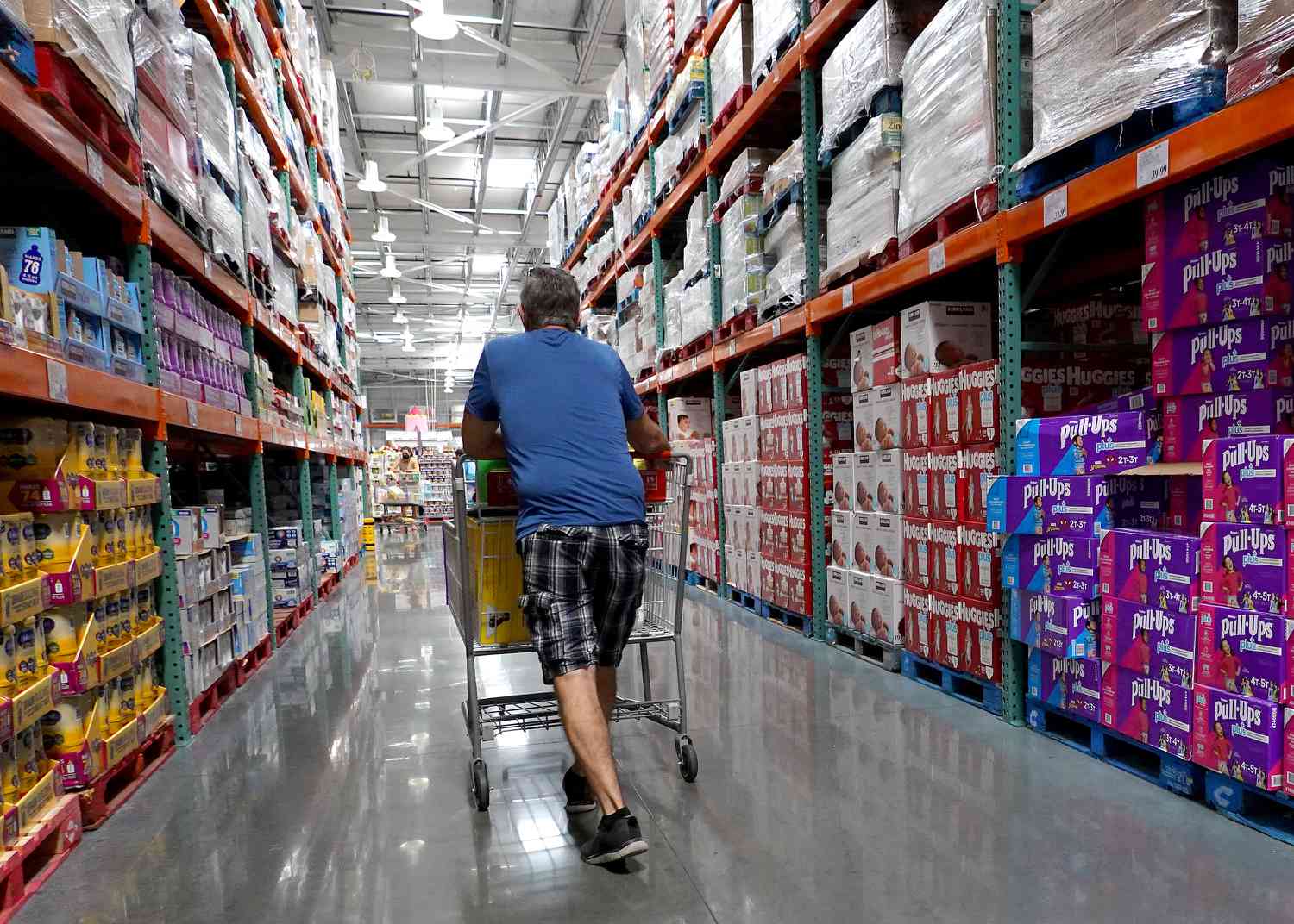 A man with his back to the camera pushes a shopping cart down an aisle at Costco.