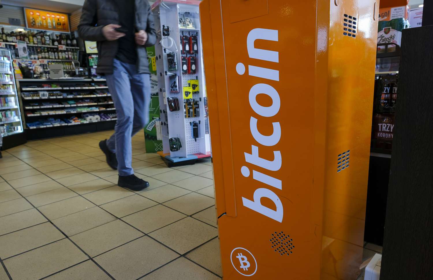 A man walks past an ATM machine in a gas station. The ATM machine has the bitcoin logo on it.