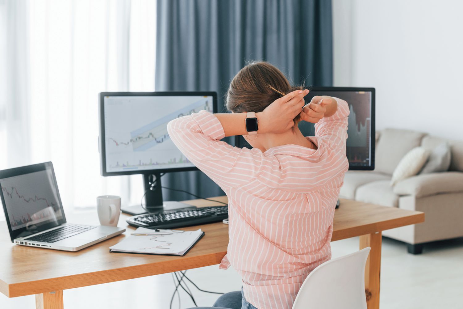 Stock trader tying hair up while following stock's chart patterns and trends on screens.