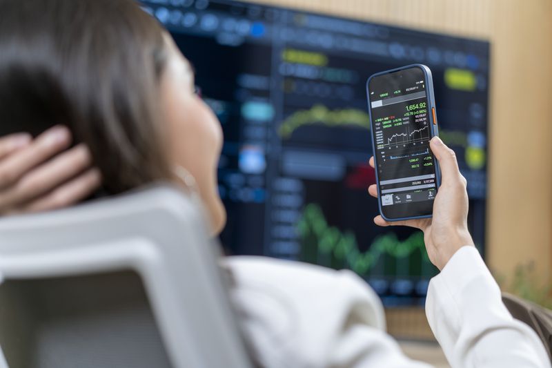 Woman looking at a stock chart on a smartphone with another larger screen with stock charts on it in the background
