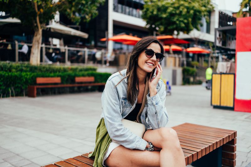 Young woman talking on the phone while sitting on a wooden bench outside.