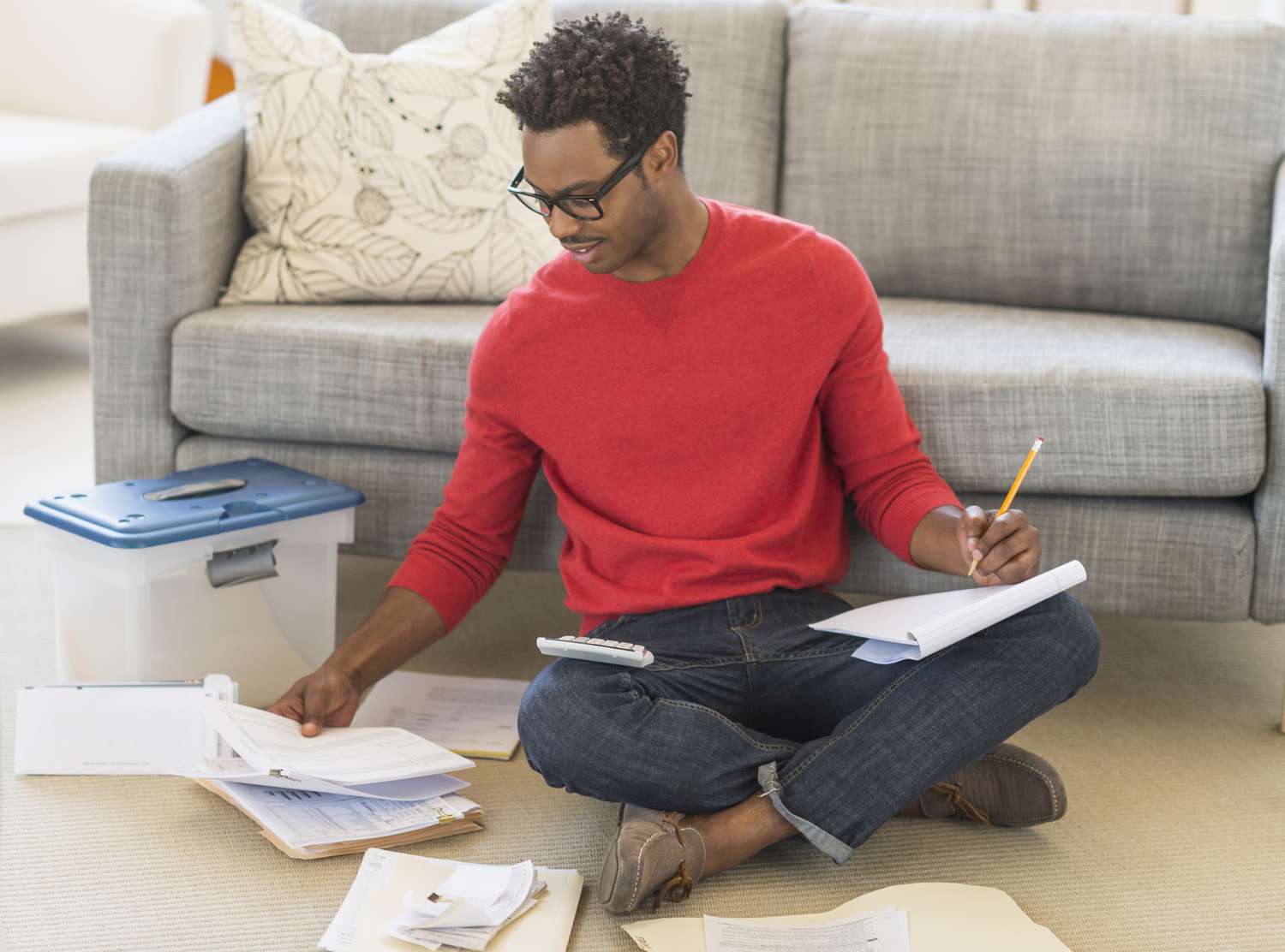 Man sitting on floor in living room working on bills and using a calculator, pen, and notepad