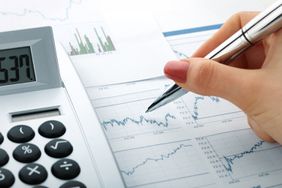 A hand holding a pen analyzing financial graphs on paper near a calculator and a rough bar chart on a document