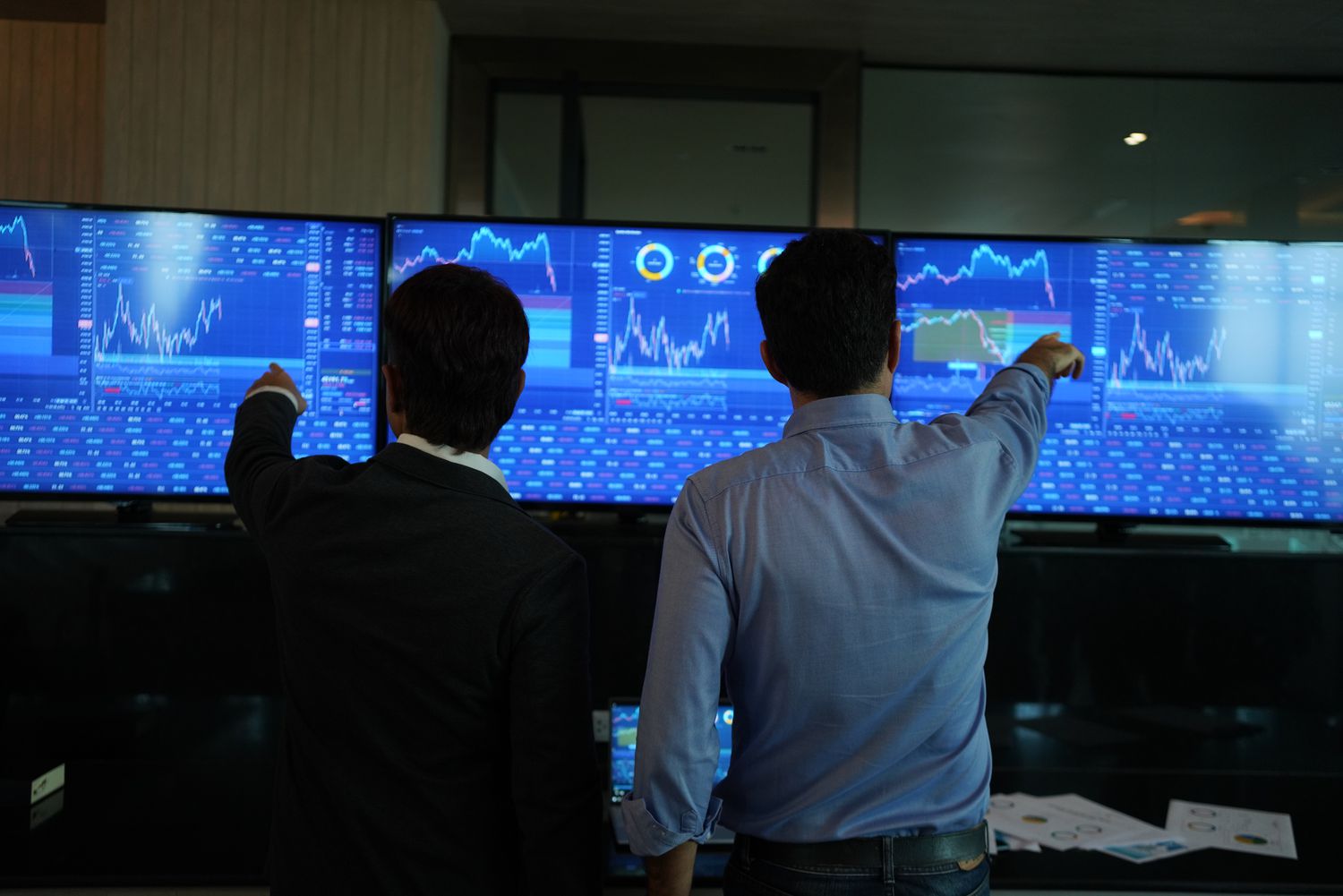 Two people pointing at a bank of stocks screens.