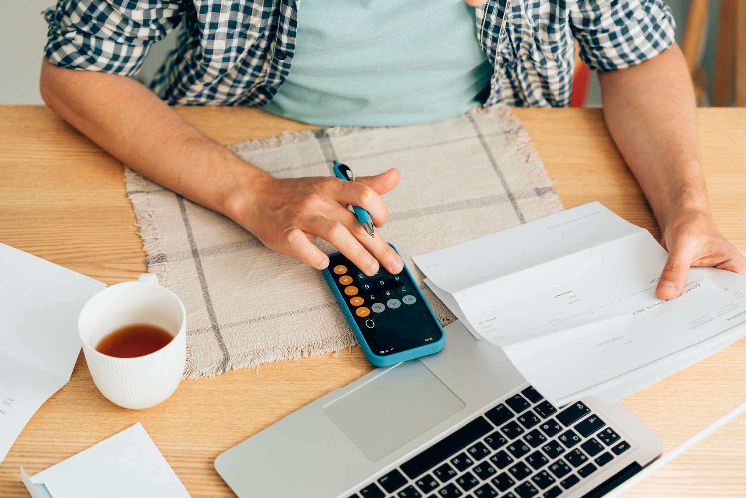 Person typing on phone calculator at table with computer, coffee, and documents arrayed