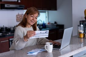 A smiling woman looks at documents and a laptop at a kitchen table.