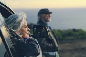 An older woman leans on a car door facing a sunset, while an older man leans against the ood