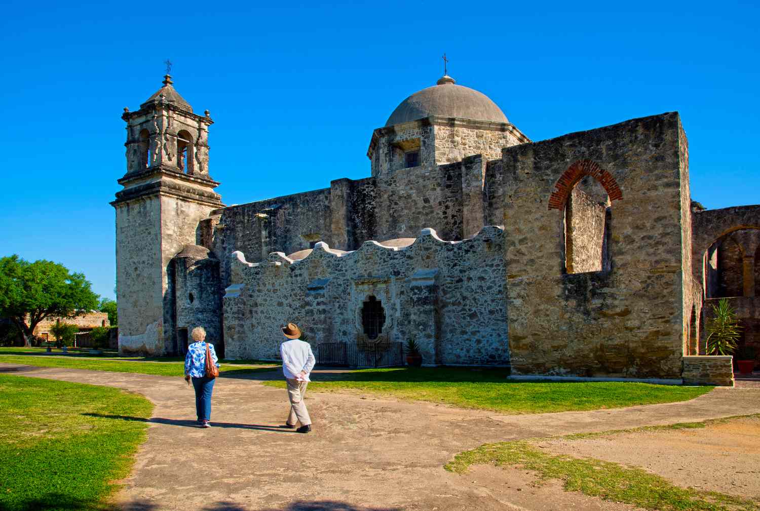 Two people walking at The Mission San Jose was founded in 1720 by Catholic religious orders to spread Christianity, The church was built in 1768 from local limestone and has become a popular tourist attraction. The Mission is now part of the San Antonio Missions National Historical Park and is designated a UNESCO World Heritage Site.